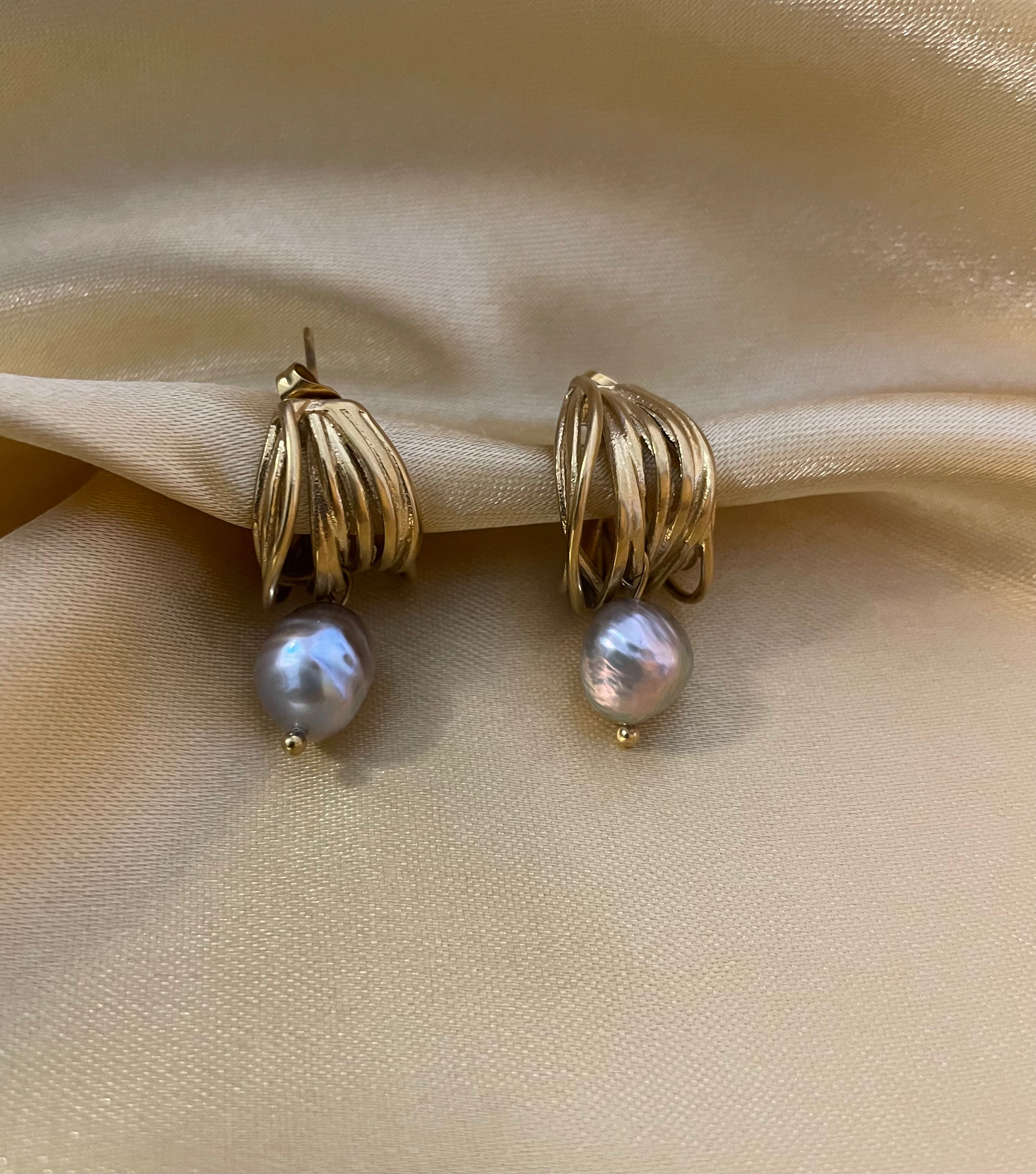 Luxury fashion jewelry photography of gold twist hoop earrings with dangling grey pearls. High-resolution close-up on the ear showing realistic skin texture, intricate gold detailing, and a sophisticated silver outfit.